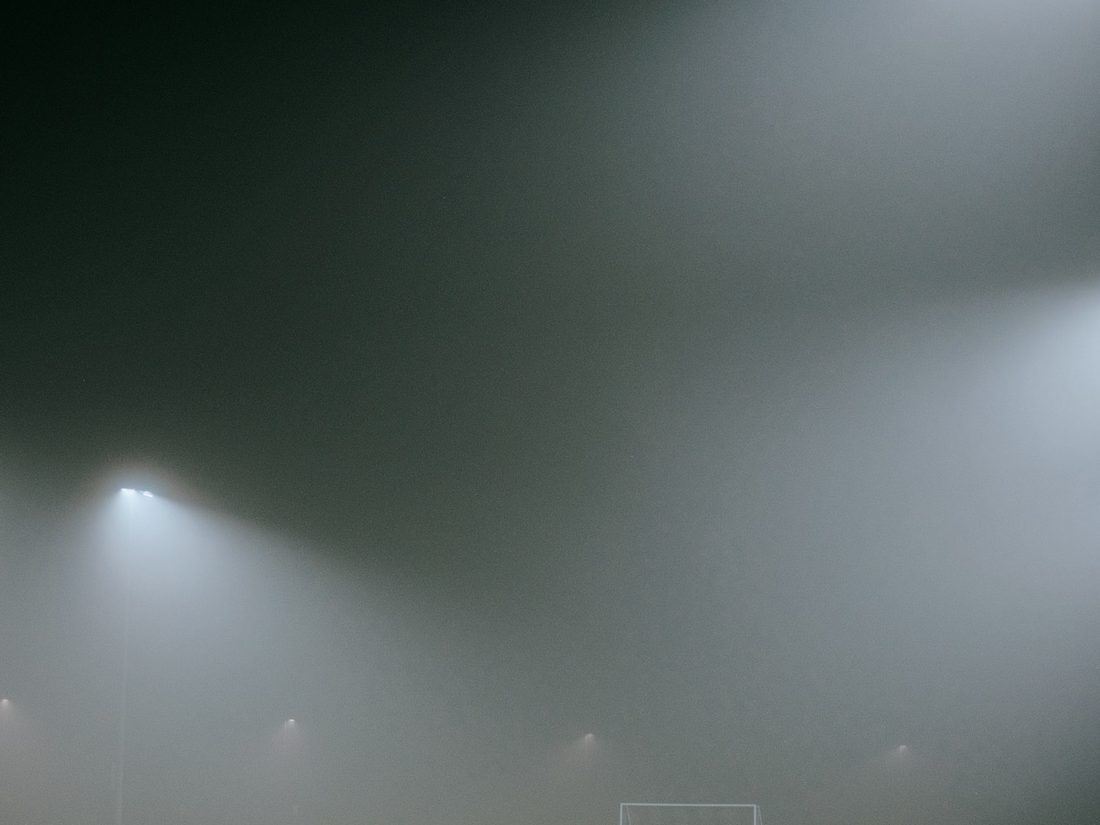 Floodlights burning through fog over an empty pitch